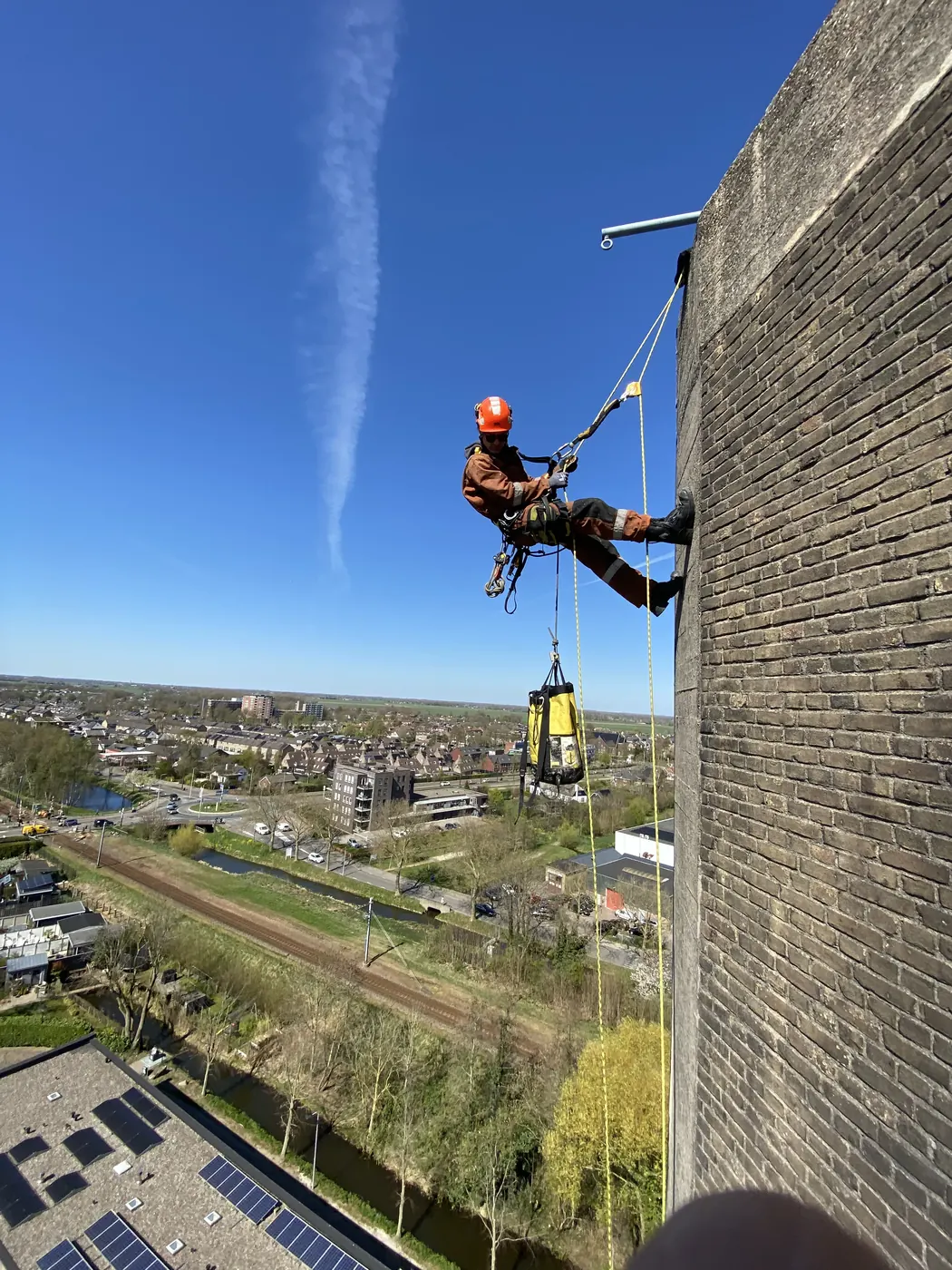 Touwtoegangstechnicus in oranje overall hangt aan de gevel van een bakstenen watertoren boven een woonwijk